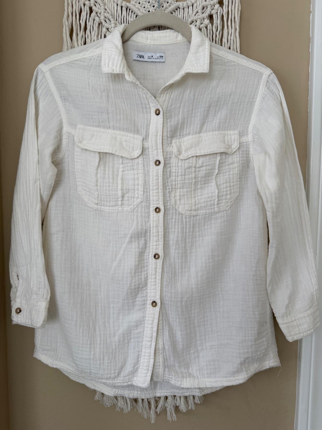 White long-sleeve shirt with front pockets and brown buttons, hanging on a hanger against a beige background with macrame.