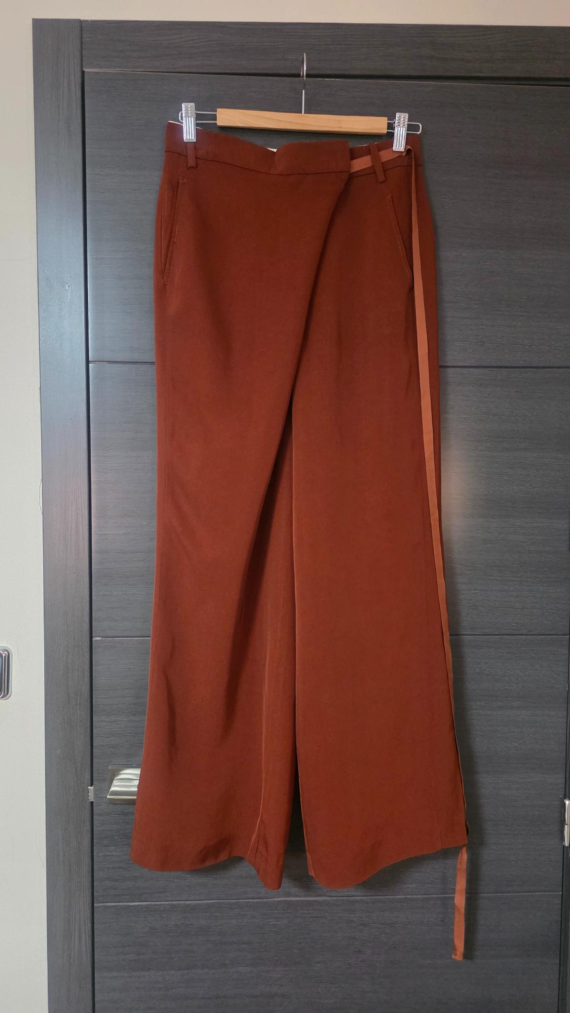 Terracotta wide-leg trousers with an asymmetric wrap front and side ribbon, hanging on a wooden hanger on a dark grey door.