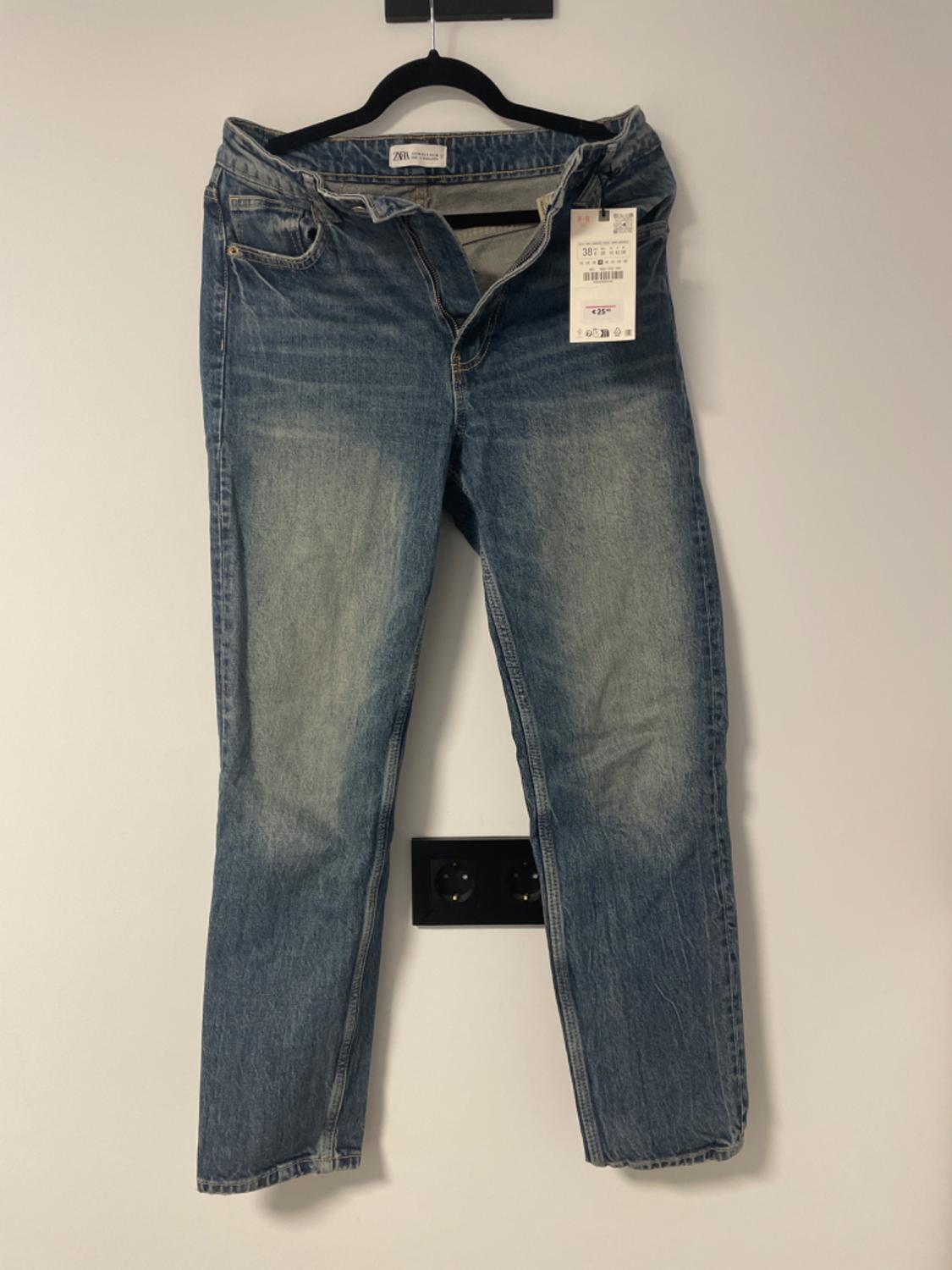 Worn blue jeans hanging on a black hanger against a white wall. Labels visible on the waistband.