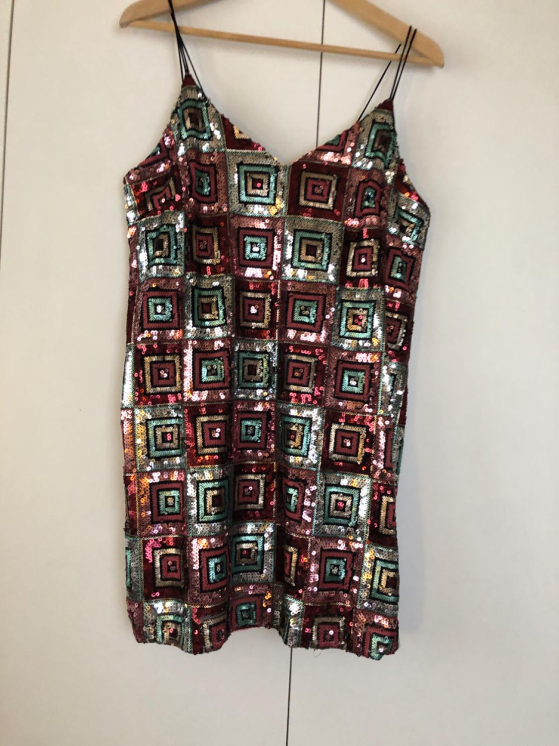 Short, spaghetti-strap dress with multi-coloured sequins forming square geometric patterns, hanging on a wooden hanger.