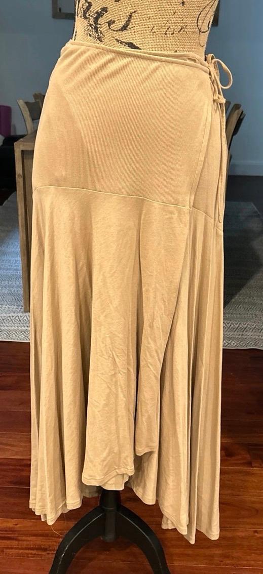 Long beige skirt on a mannequin indoors. It has a wrap design with a side tie, a ribbed upper section and an asymmetrical, flowing hem.