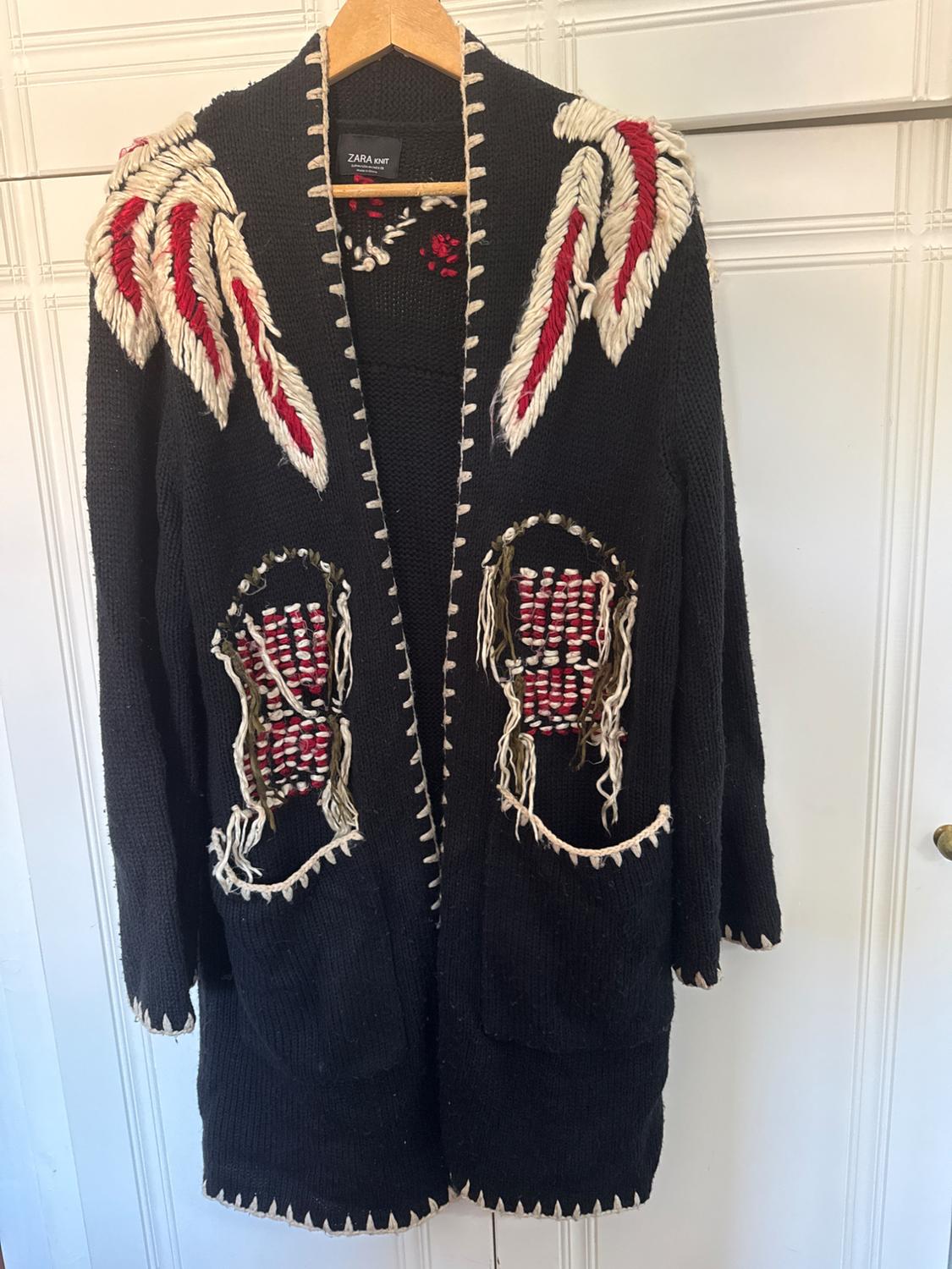Black cardigan hanging on a hanger, with knitted red and white details on the shoulders and pockets. Visible label: ZARA Knit.
