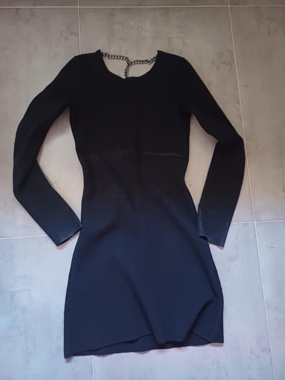 Short, tight-fitting, long-sleeved black dress with chain detail at the neck, laid out on a grey tiled floor.