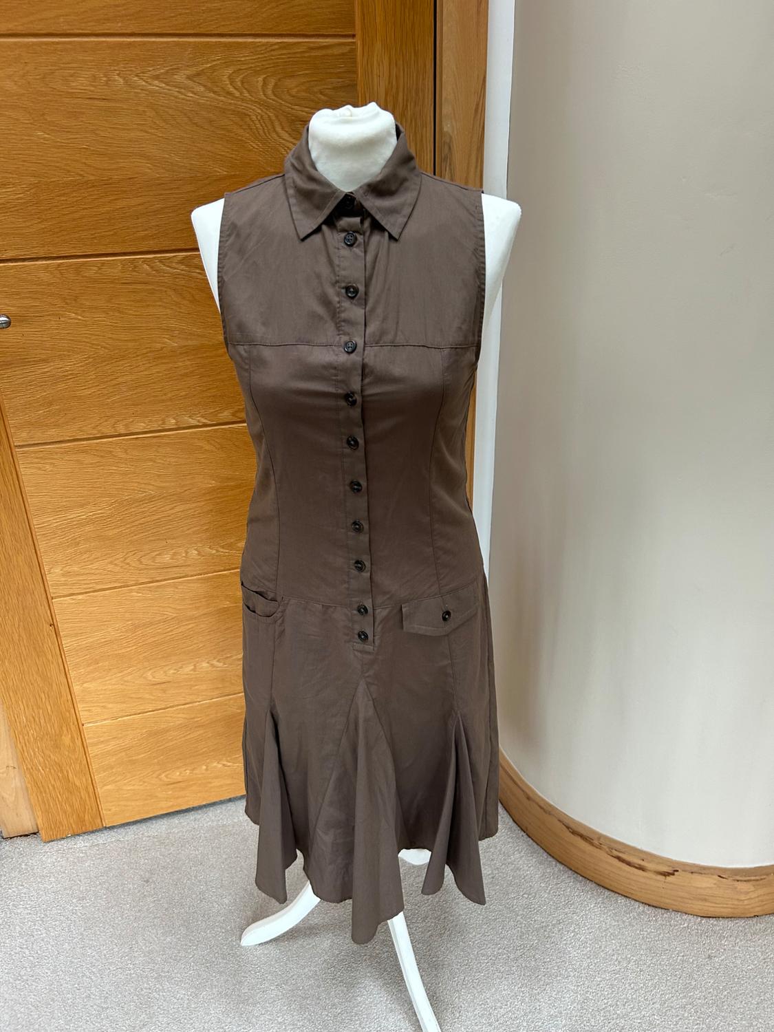 White mannequin wearing a sleeveless brown dress with buttons, a collar and pleated skirt, in an interior with a wooden door.