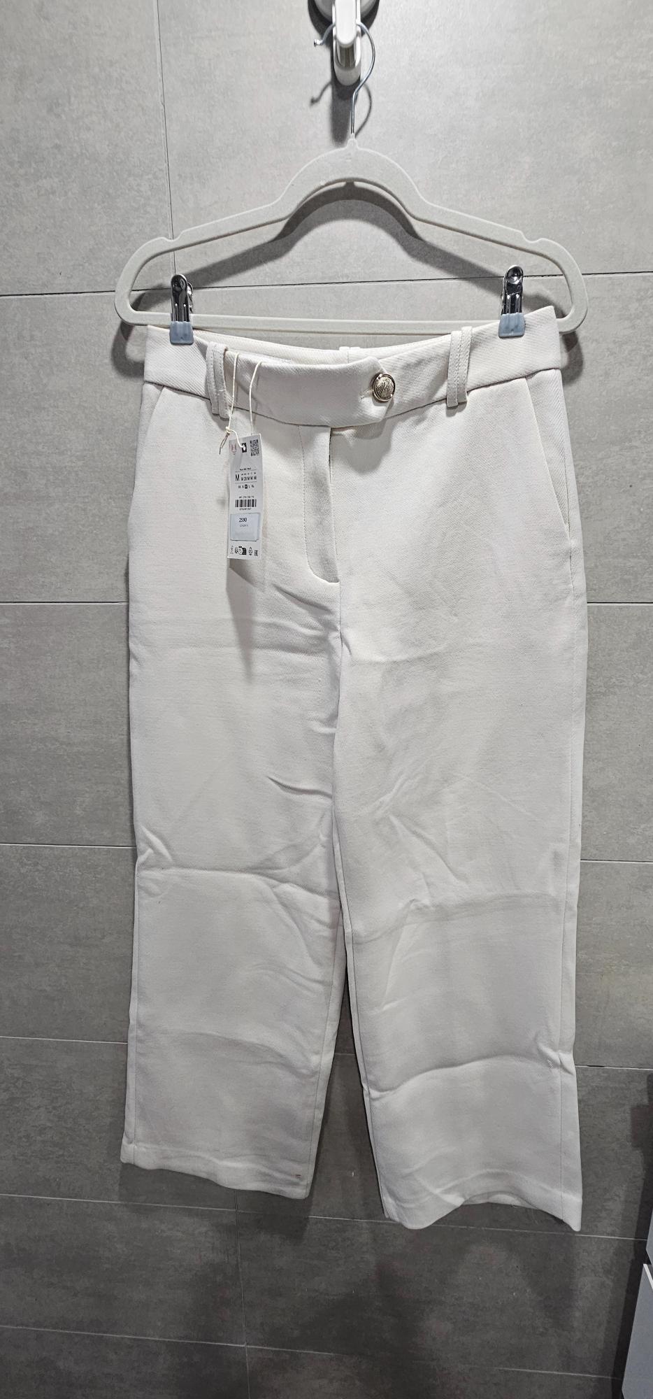 White trousers hanging on a clip hanger against a grey tiled wall. Shows a gold front button and a label.