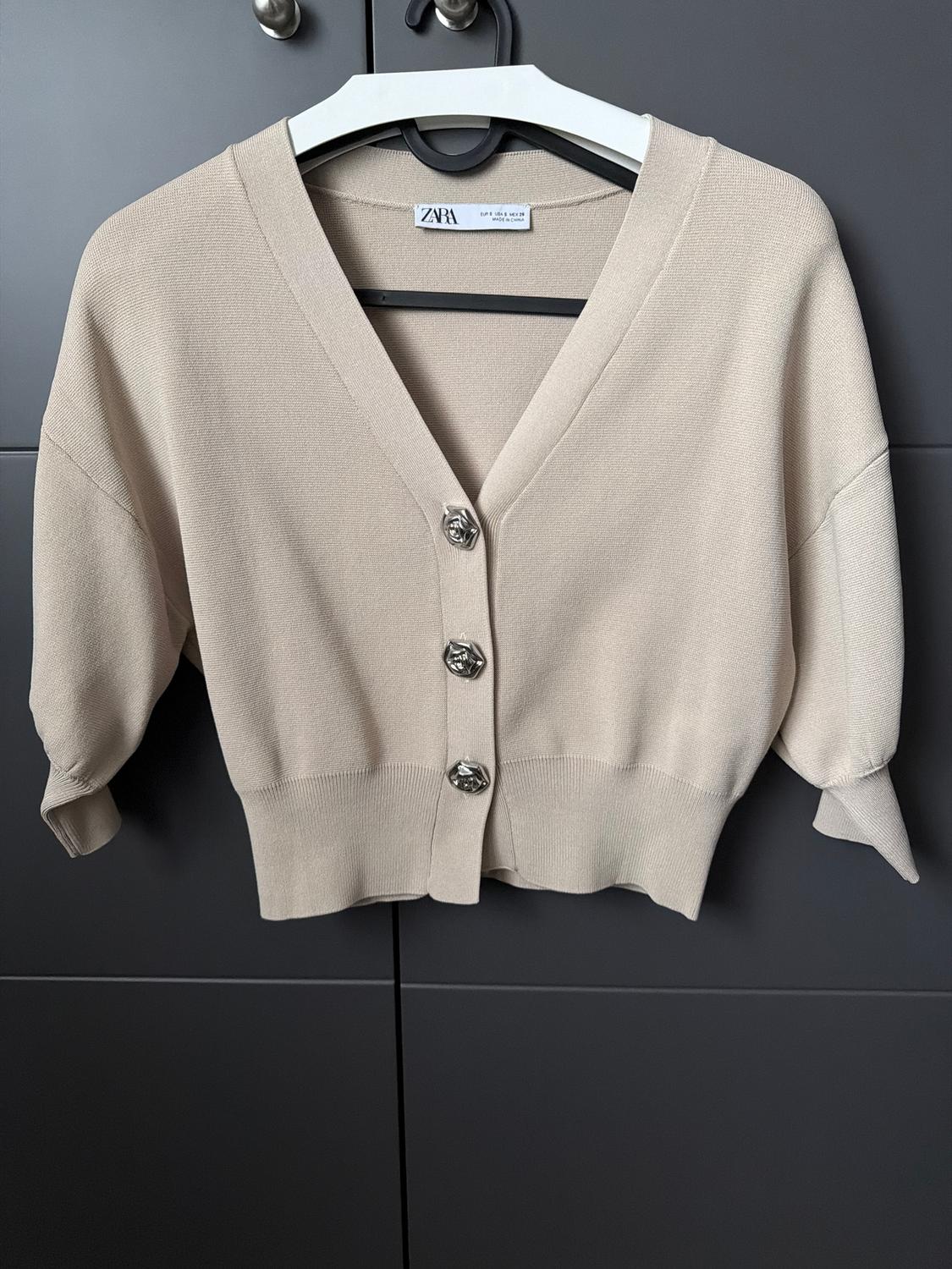 Beige short-sleeved cardigan with silver buttons, hanging on a white hanger in front of a dark grey background.