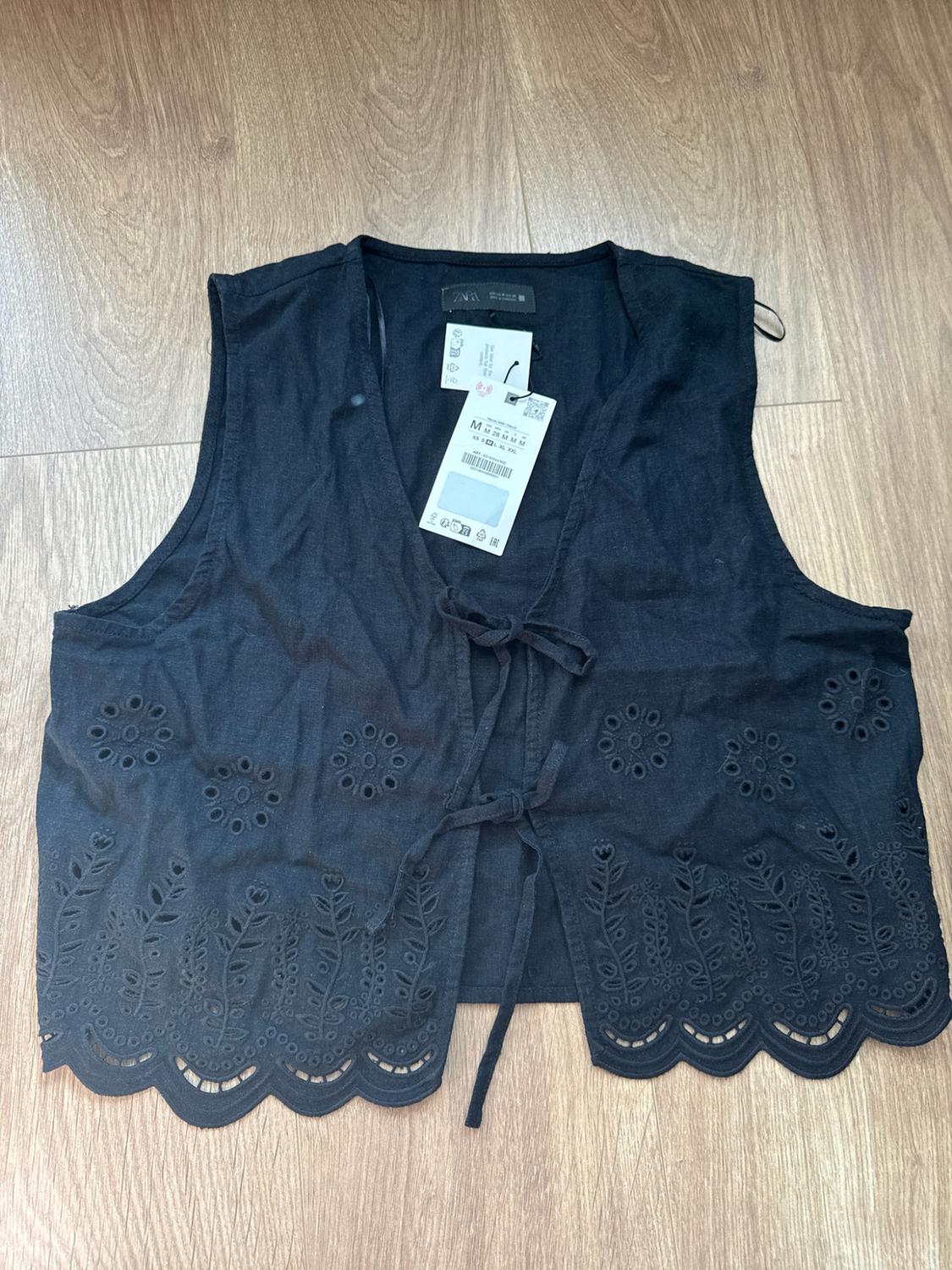 Black waistcoat on wooden floor. Design with perforated embroidery,
scalloped hem and front tie fastening. ZARA labels and M size visible.
