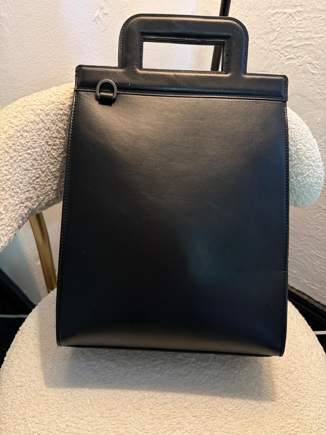 Structured black handbag with a rectangular design and integrated handle, resting on a textured white chair in an indoor setting.