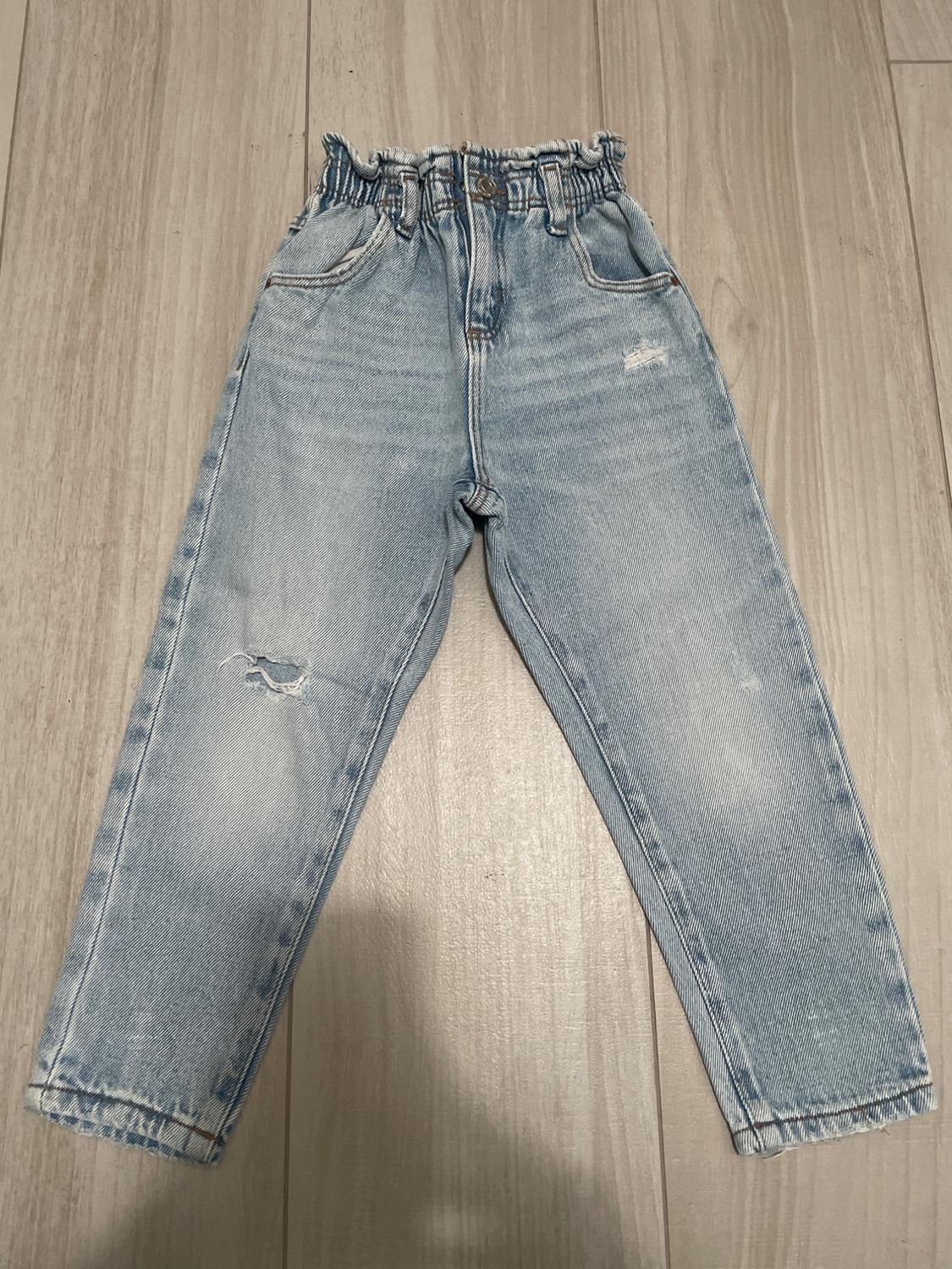 Light blue denim jeans with a shirred elasticated waist and distressed details, on a light wooden floor.