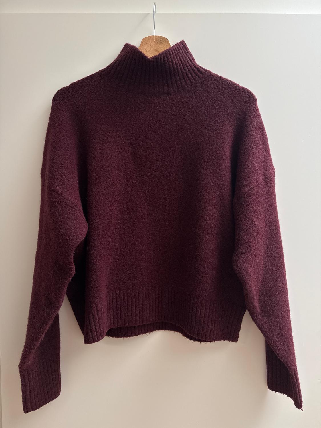 Burgundy roll-neck jumper hanging on a wooden hanger against a white background. Chunky knit design with long sleeves.