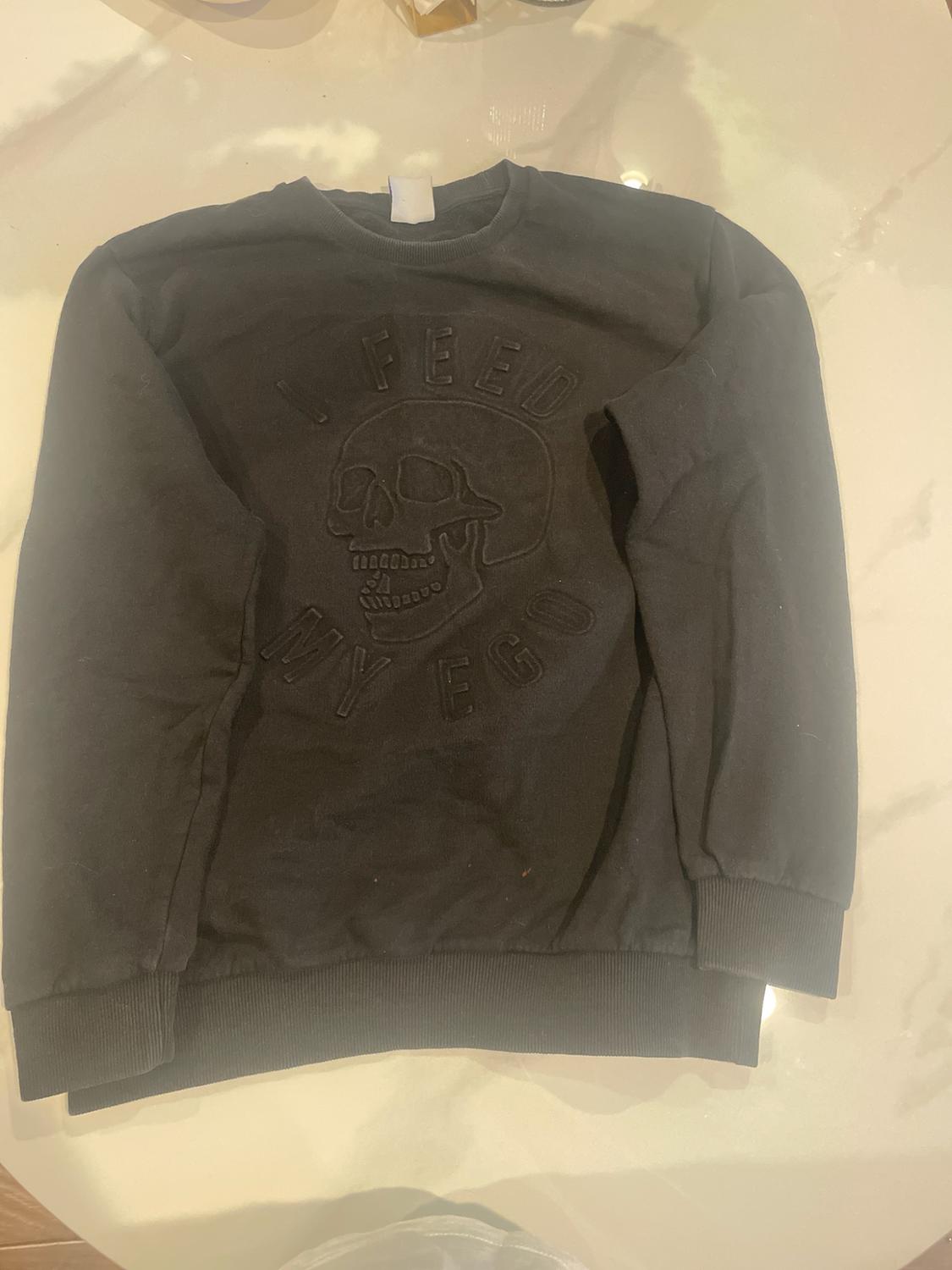Black long-sleeve sweatshirt on a light background. It features an embossed skull design
with the text I FEED MY EGO.