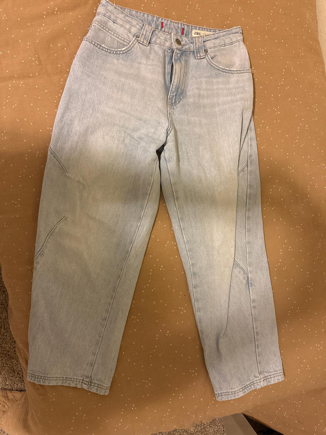 Light blue denim jeans on a brown bedspread with small white dots. The inside label reads "ZARA".