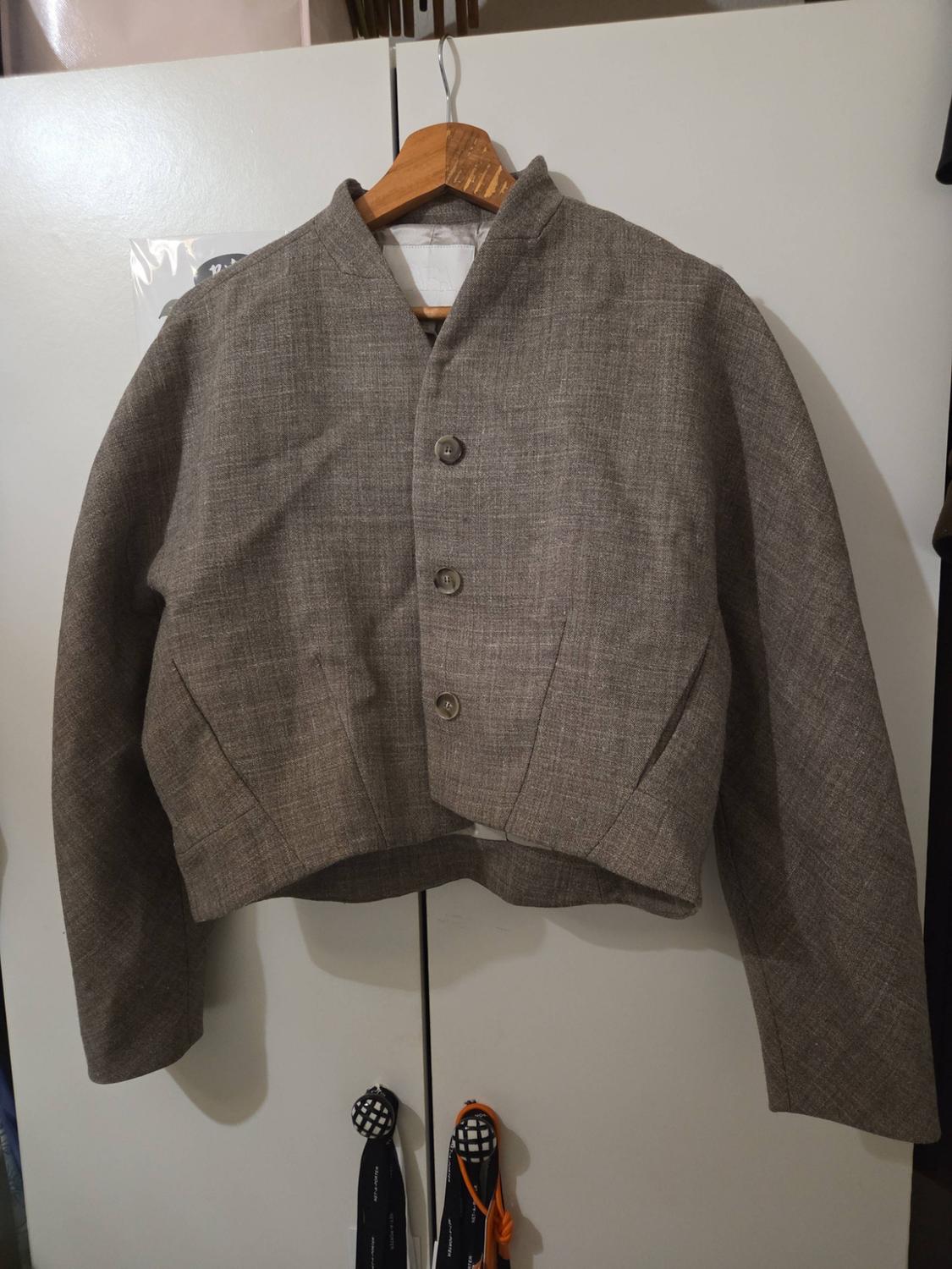 Grey textured cropped jacket, three buttons and long sleeves, hanging on a wooden hanger above a white wardrobe.