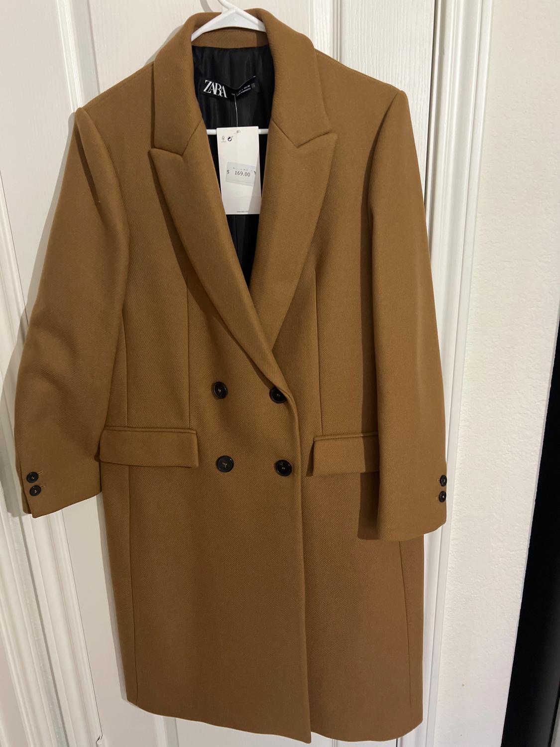 Double-breasted camel coat hanging on a white door. Visible tag with price of $169.00.