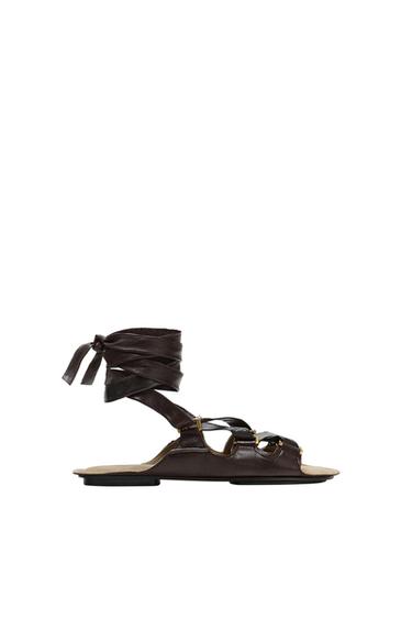 Dark brown flat sandal with crossed straps to tie around the ankle and gold-tone metal details on a white background.