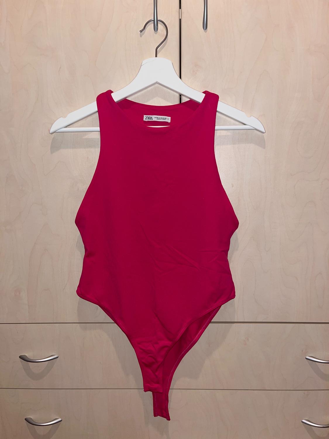 Sleeveless red bodysuit hanging on a white hanger, light wood background with metal drawers visible at the bottom.