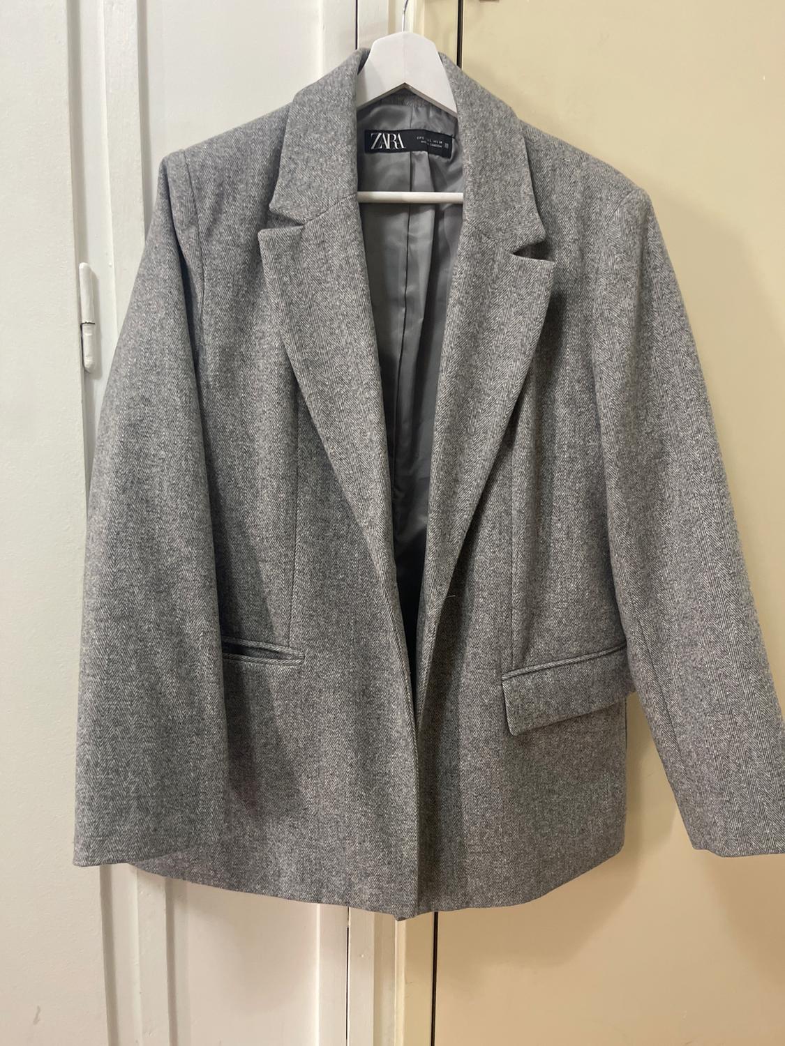 Grey wool jacket hanging on a white hanger, with wide lapels and front pockets. Visible label inside.