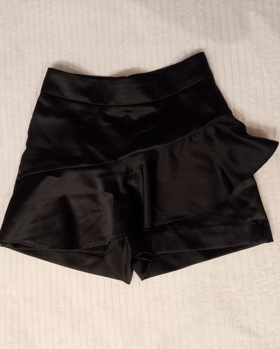 Black satin-finish skort with asymmetric frill, photographed on a white textured surface.
