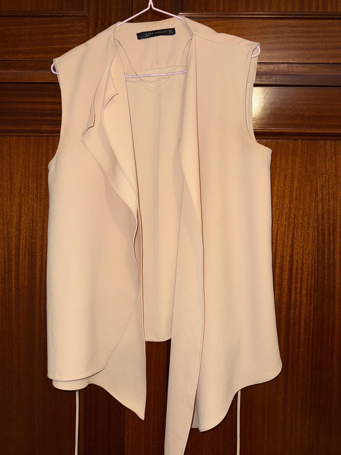 Sleeveless beige waistcoat hanging on a white hanger over a wooden door. Visible label: "Zara Woman".