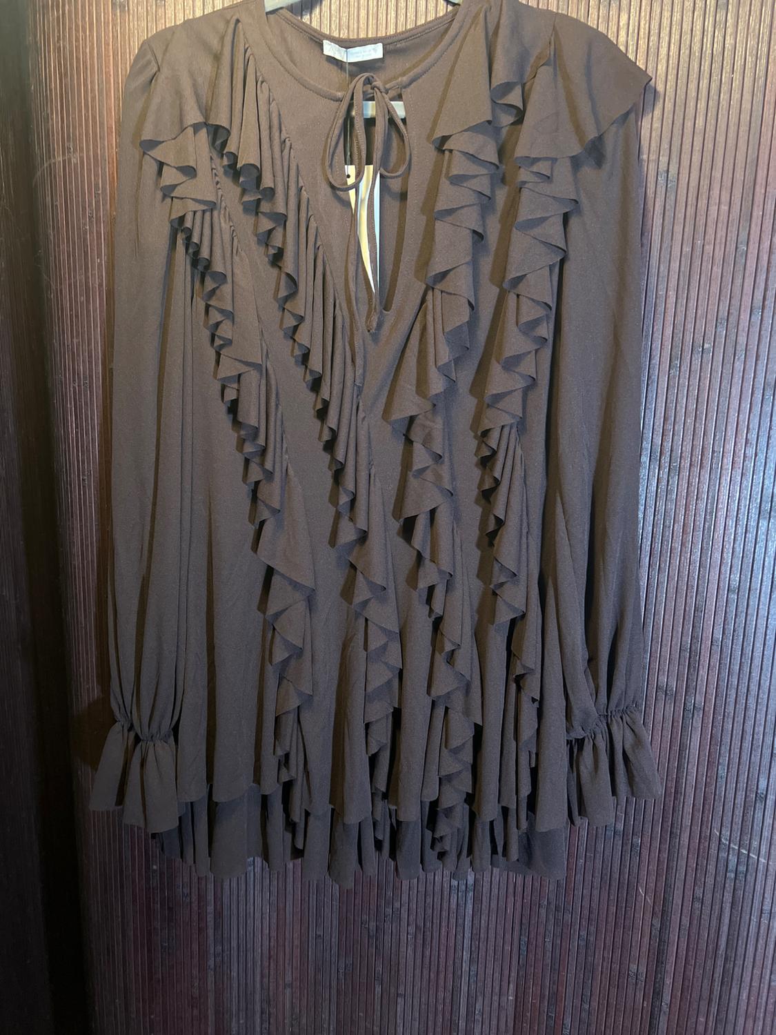 Brown dress with cascading ruffles, long sleeves, and a bow at the neck, hanging on a textured wooden wall.