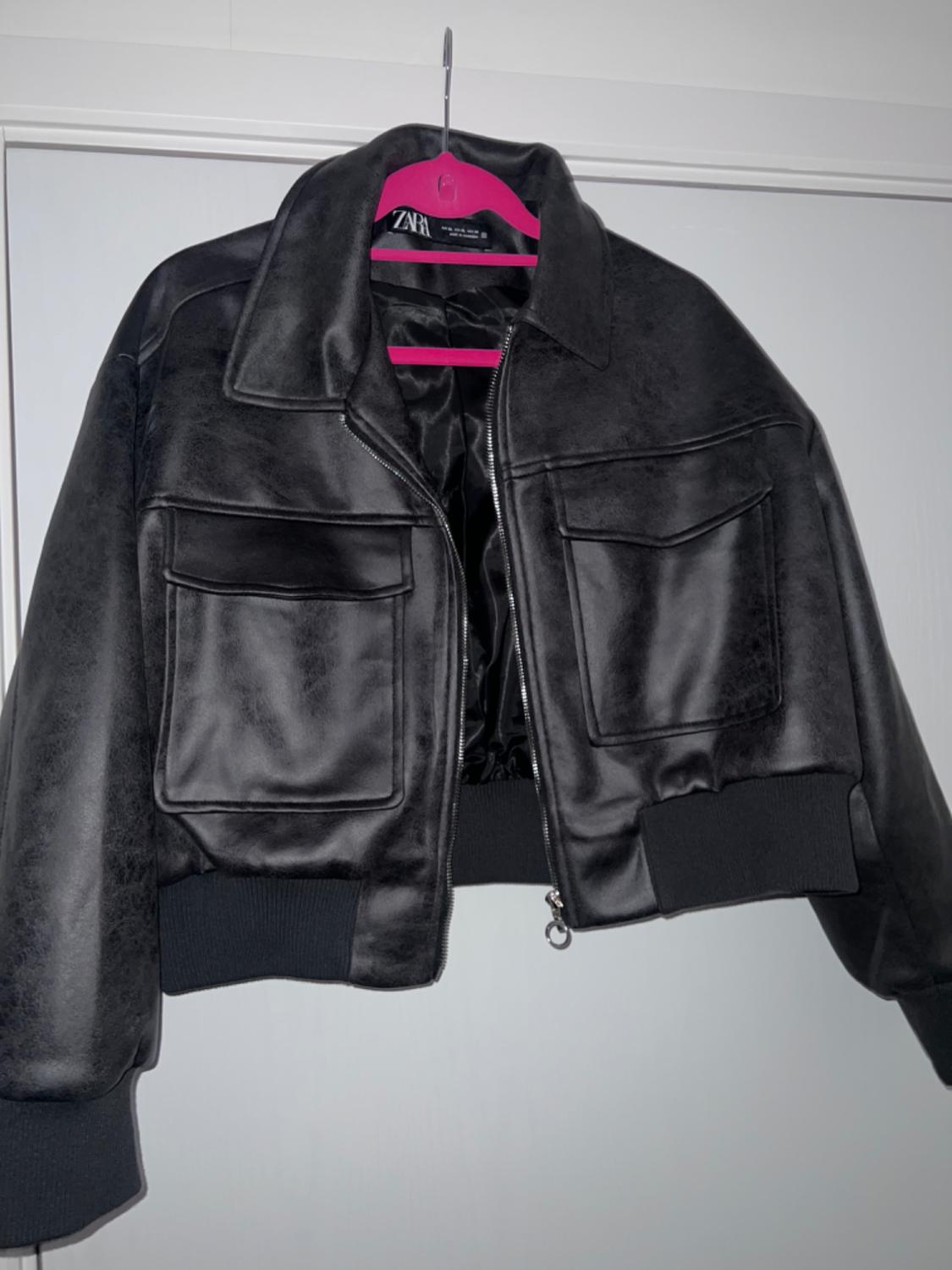 Black faux leather jacket hanging on a pink hanger. It has large pockets and a front zip fastening. Visible label: ZARA.