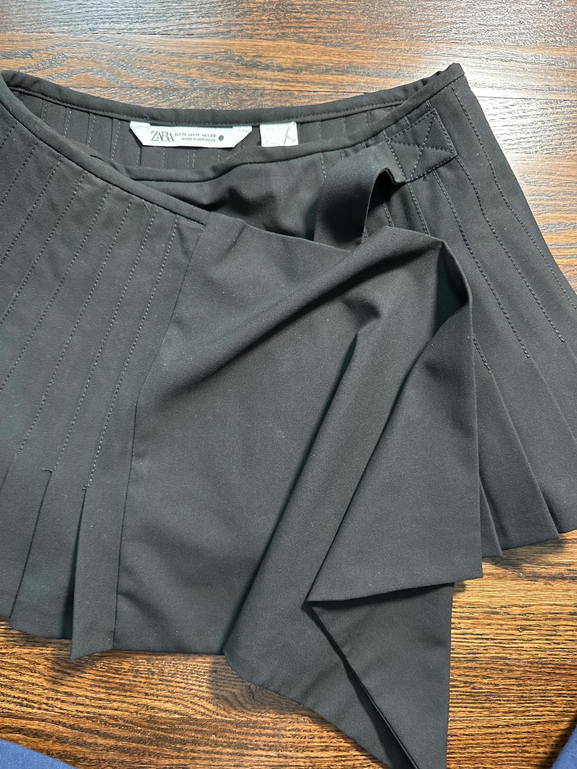 Black skirt with pleats and draped detail on a wooden surface. Visible label with text: "ZARA".