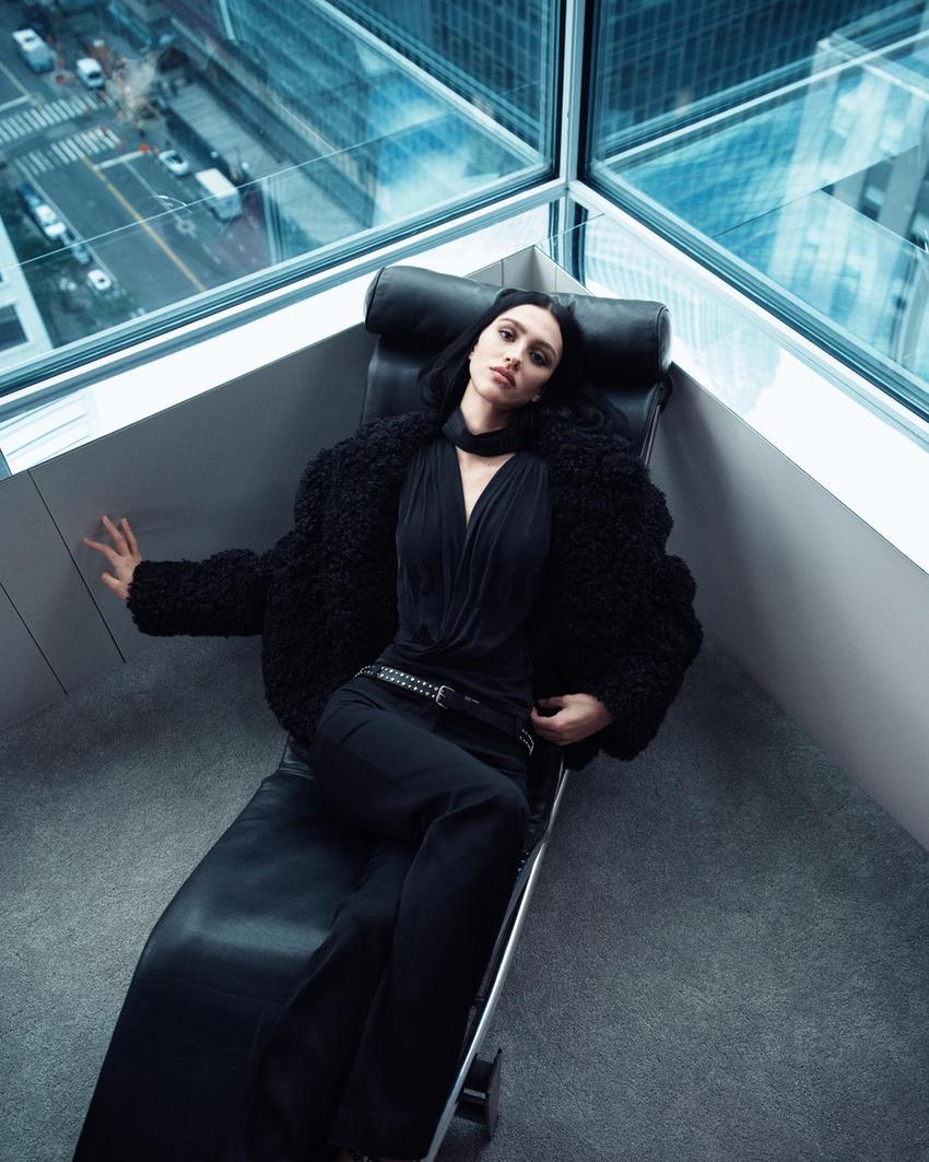 ZARA x Ludovic de Saint Sernin: Kolekcija koja definira novu estetiku senzualnosti 1 Woman in a black fuzzy coat and dark pants, reclining in a modern chair by a window with a city view.