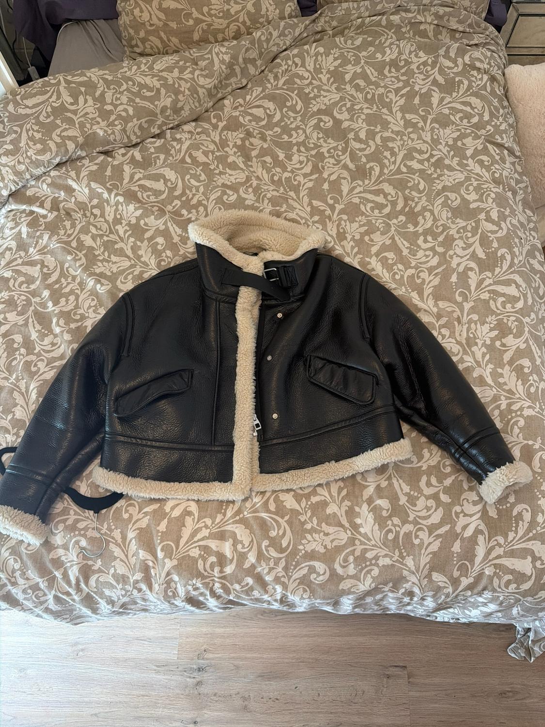 Black jacket with a light-coloured fur lining and a buckle at the neck, placed on a bed with a beige floral-patterned bedspread.