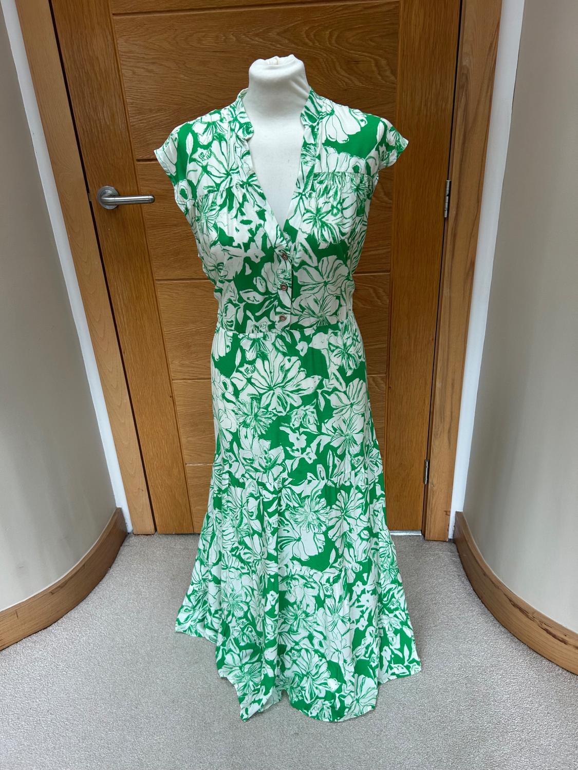 Mannequin in a long green dress with a white floral print, short sleeves and a button-up V-neck, in front of a wooden door.