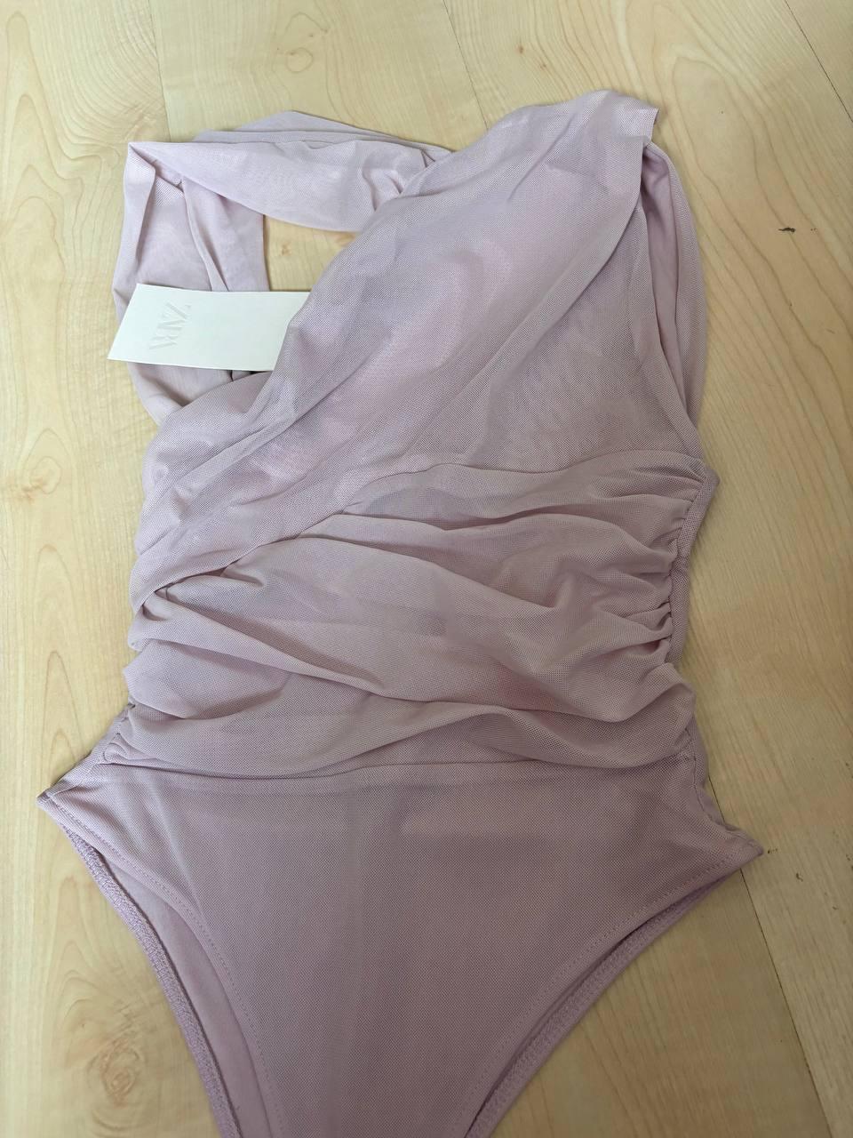 Light purple soft fabric bodysuit on a light wood background. Visible white label with "ZARA" text.