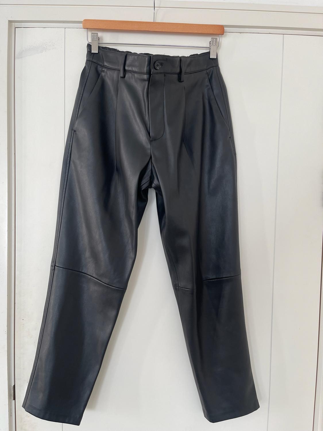Black trousers hanging on a wooden hanger in front of a white door. Design with a button fastening, side pockets and stitching.