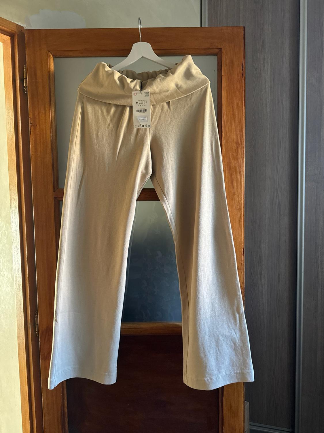 Wide beige trousers with a folded waistband and label, hanging on a white hanger on a wooden door with glass.