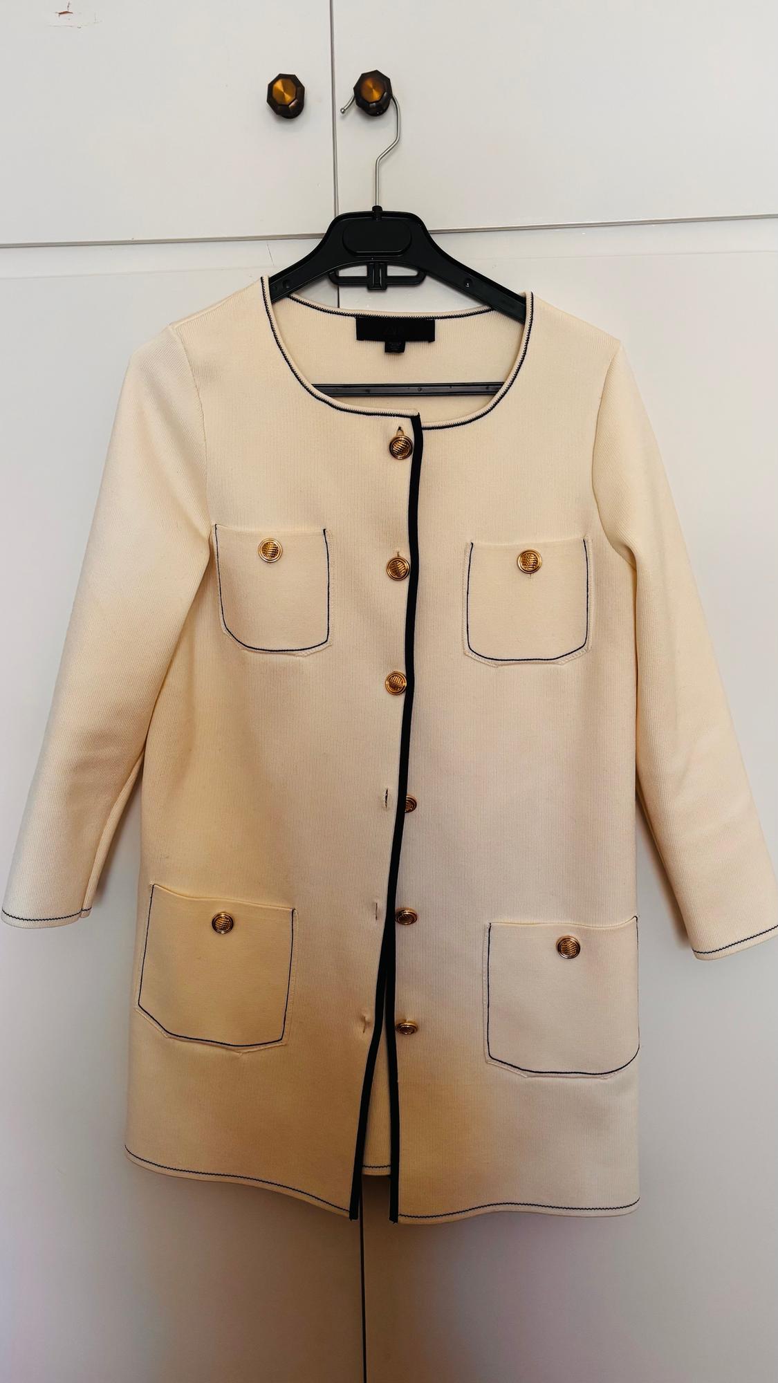 Beige jacket with gold buttons and black trim, hanging on a black hanger against a white background.