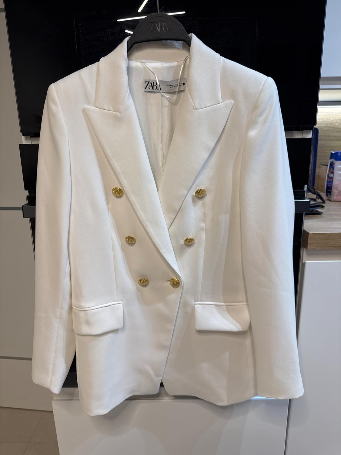 White double-breasted jacket with gold buttons, hanging on a black hanger with the brand visible on the label.