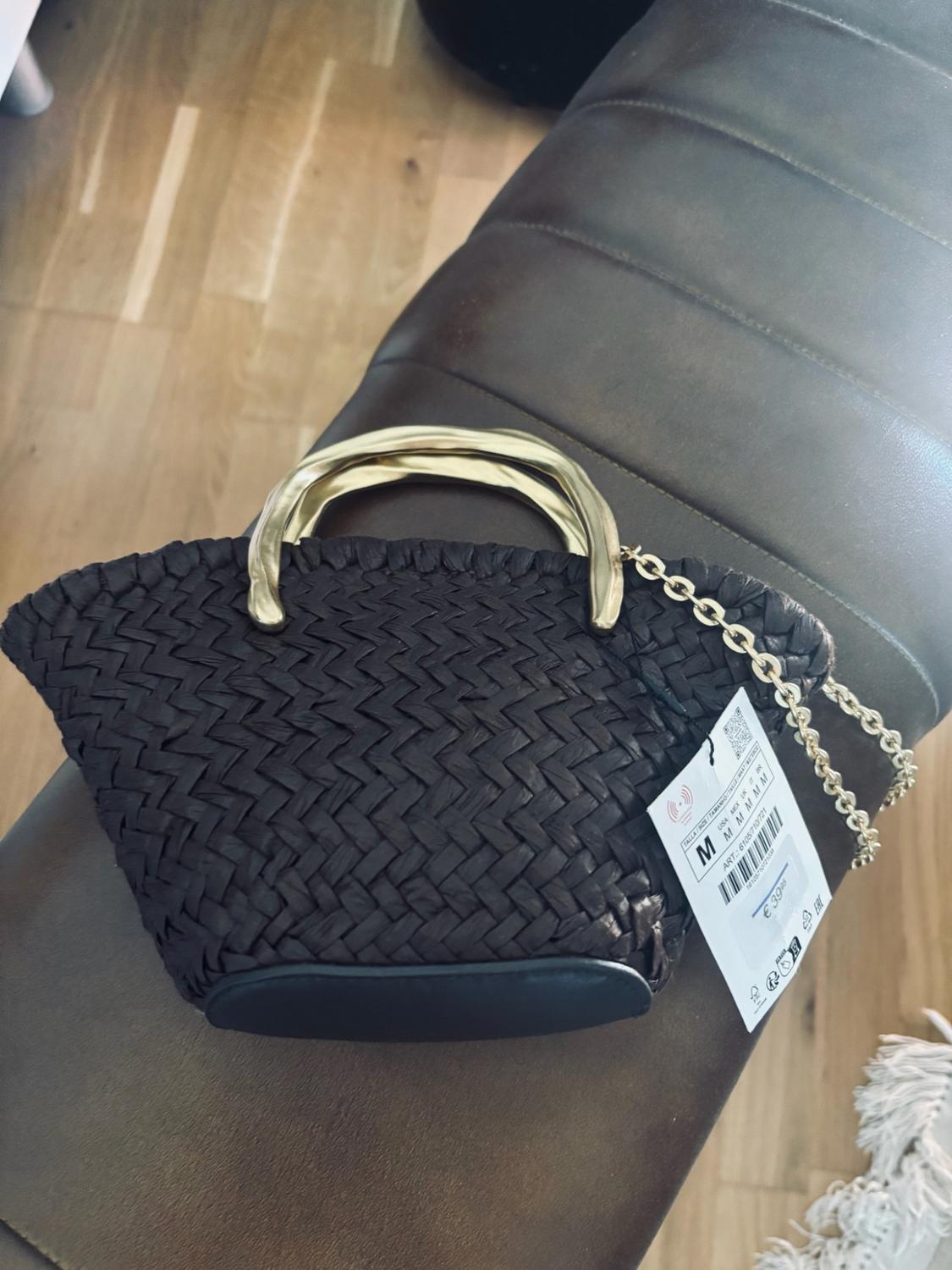 Dark braided bag with a gold handle and chain on a leather surface. Label with text: M ART: 6105/710/721 €39.95.