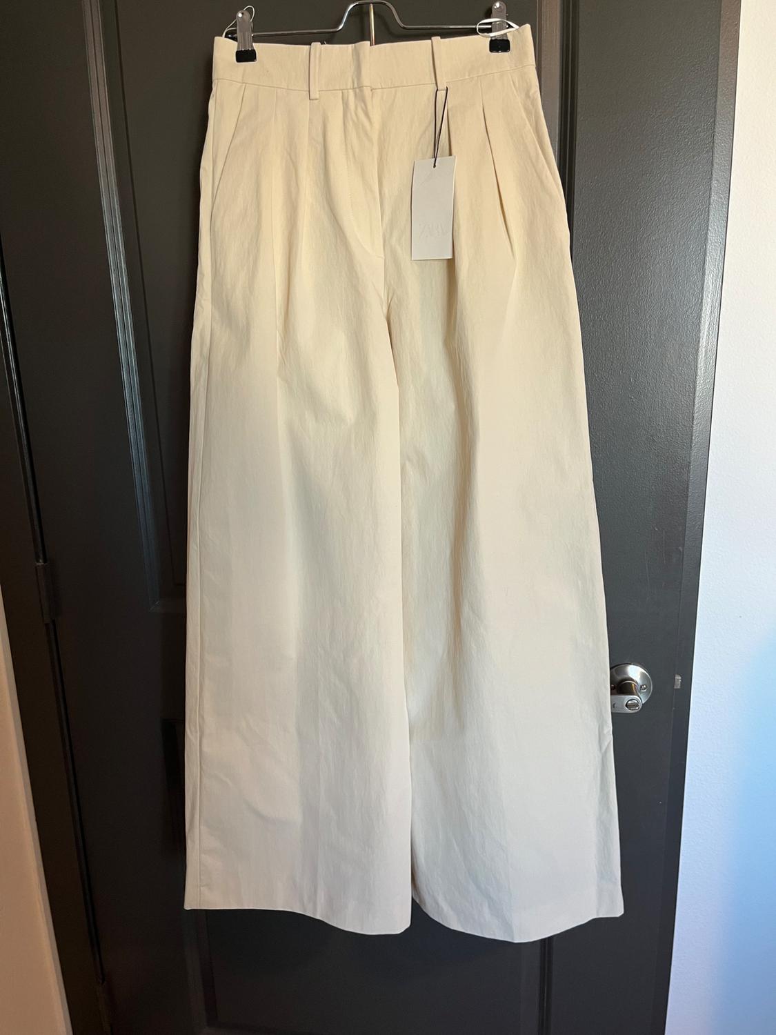 Beige wide-leg pants hanging on a dark door, with a white tag visible at the waist.
