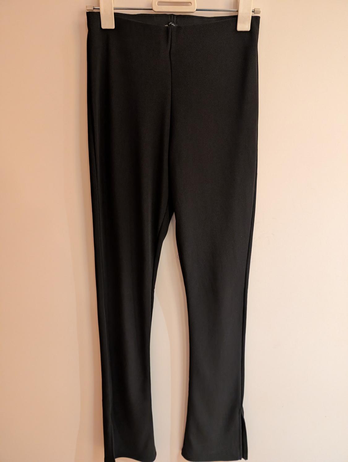 Black trousers with slits at the hem, hanging from a wooden clip hanger against a light-coloured wall. Minimalist style.