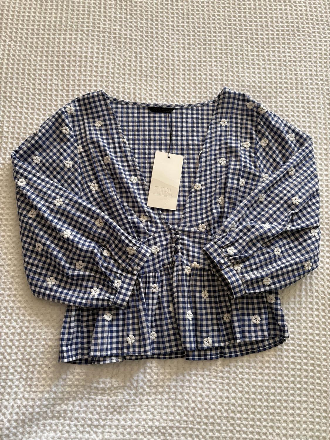 Blue and white vichy plaid blouse with small embroidered flowers, V-neck, three-quarter sleeves, and a Zara label on a white background.