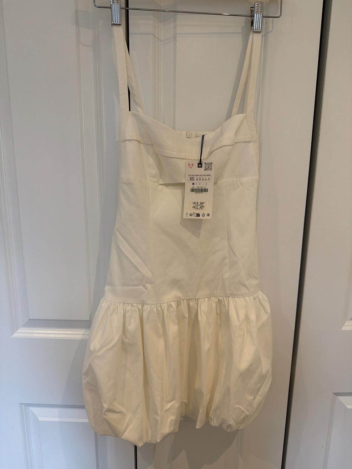 Short white spaghetti strap dress with a puff skirt hanging on a white door. Label text: XS US $ 69.90 UK £ 49.99.