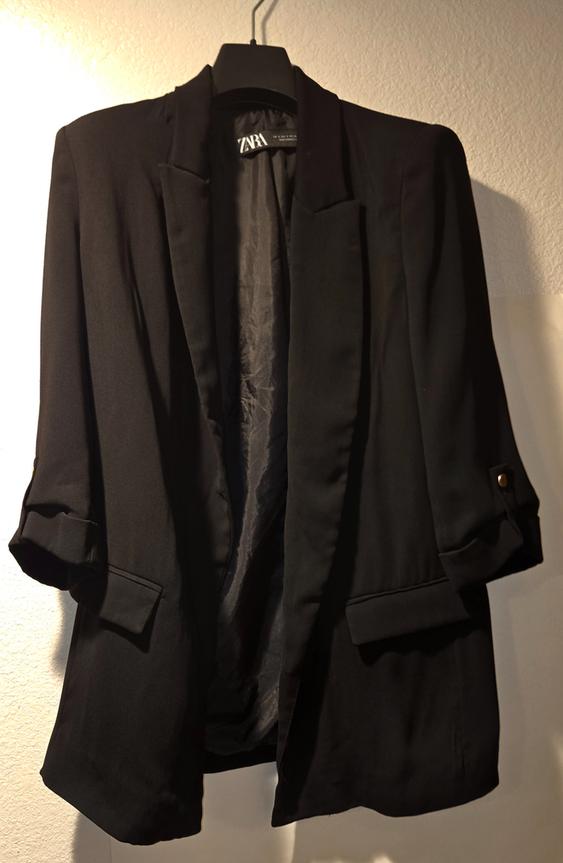 Black jacket hanging on a hanger, with wide lapels and rolled-up sleeves. Visible tag with text "ZARA". Light background.