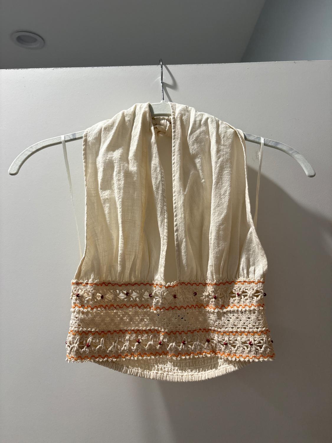 Cream sleeveless blouse with orange embroidery and floral details, hanging on a white hanger against a gray background.