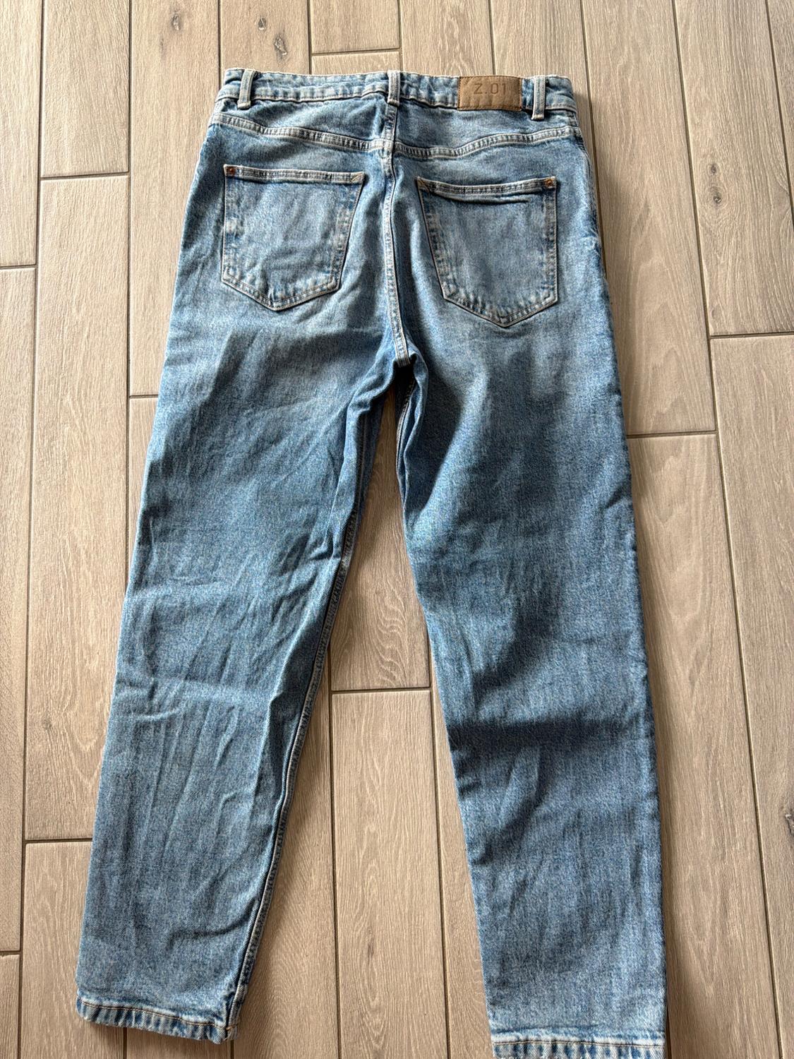 Blue denim trousers viewed from the back on a wooden floor. A brown label on the waistband featuring the text Z.01 stands out.