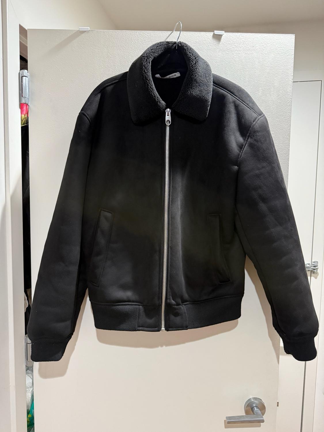 Casual style black jacket with silver zipper, textured collar
and elastic cuffs, hanging on a white door.