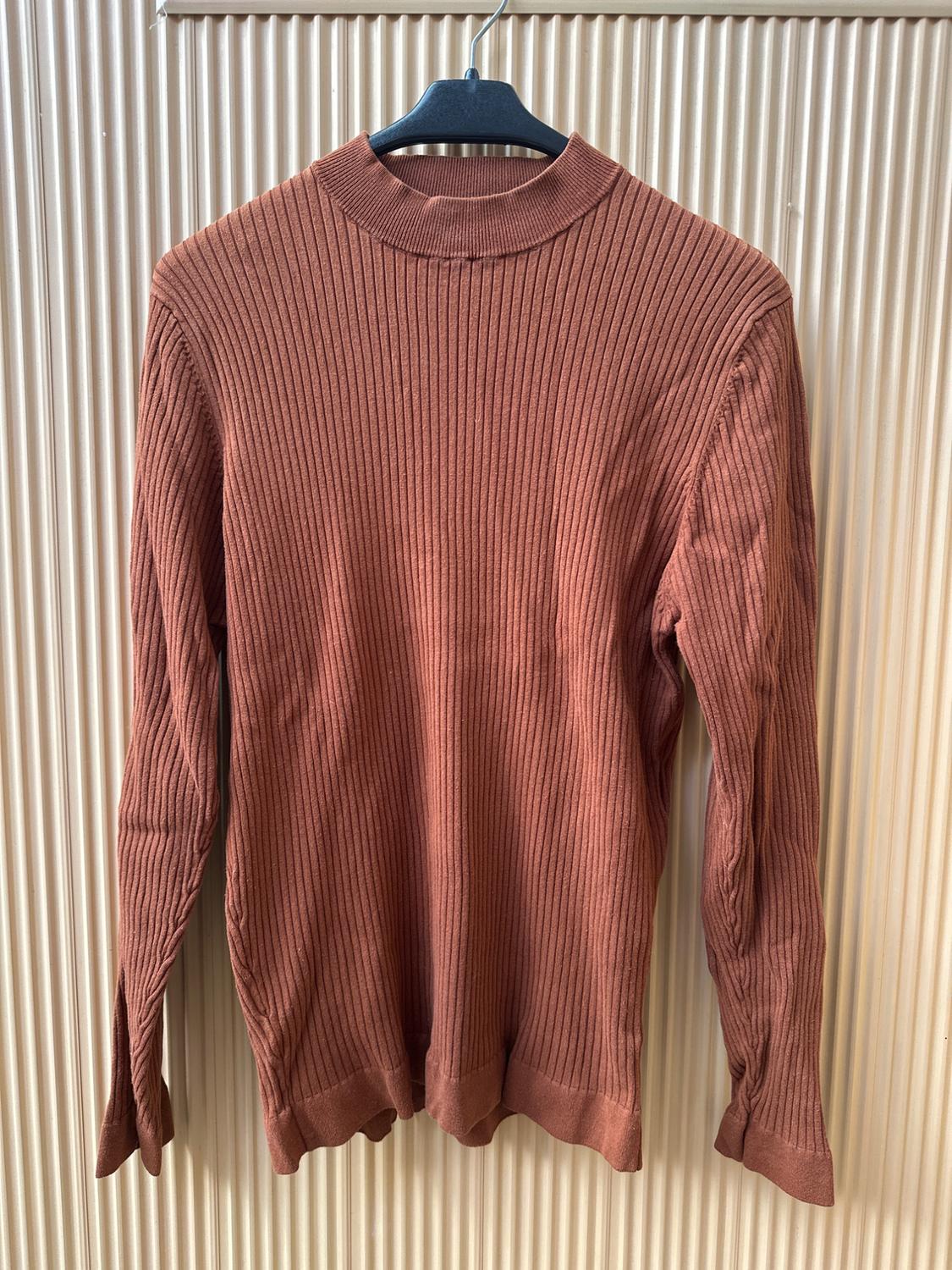 Brown ribbed knit jumper hanging on a black hanger, against a background of vertically textured beige panels.