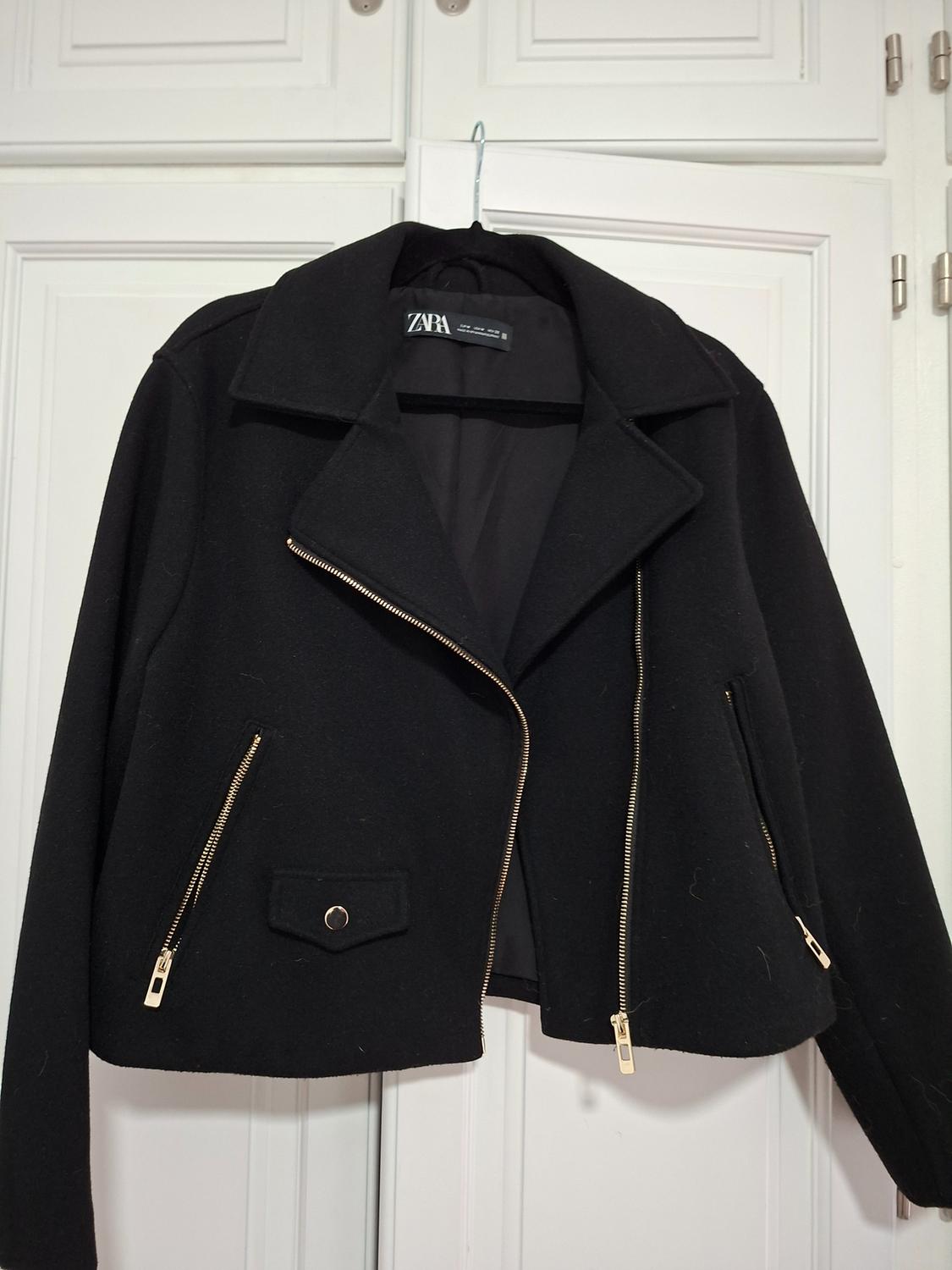 Black jacket with gold zips hanging on a white door. Visible label with text "ZARA".