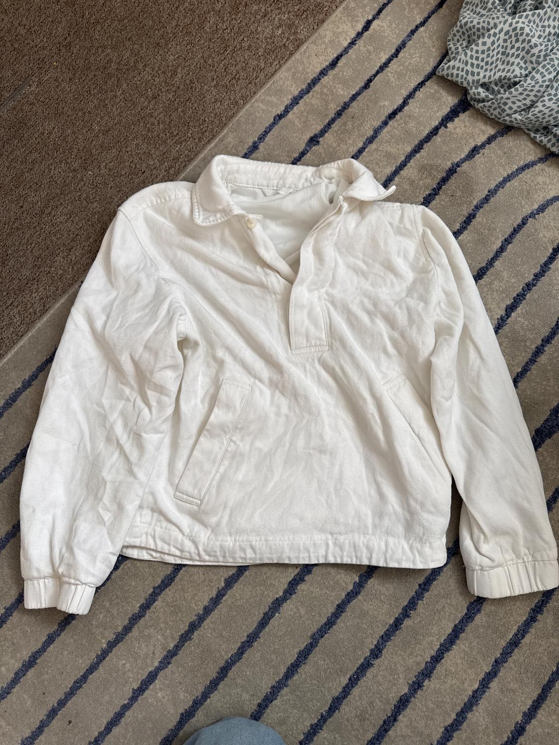 White long-sleeved sweatshirt with collar, button, and pockets, in a casual style, on a grey rug with blue stripes.