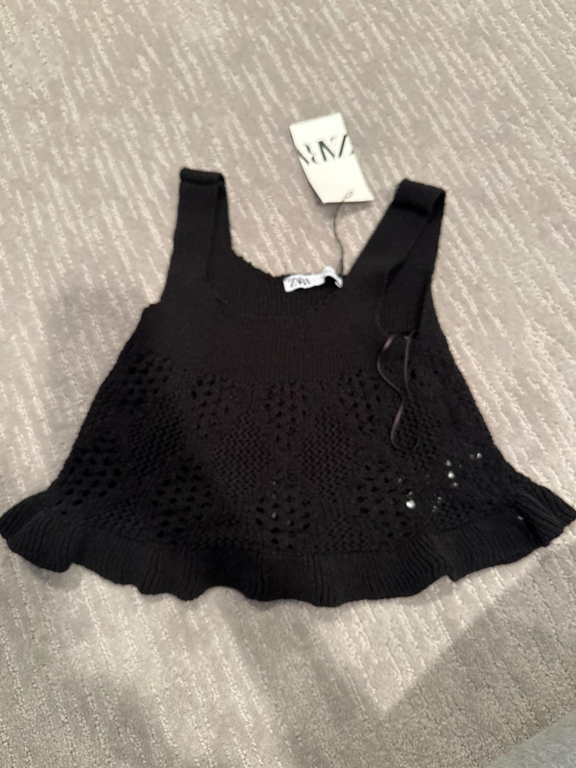 Black knit top with wide straps and openwork design on a gray rug. Visible label with text "ZARA".
