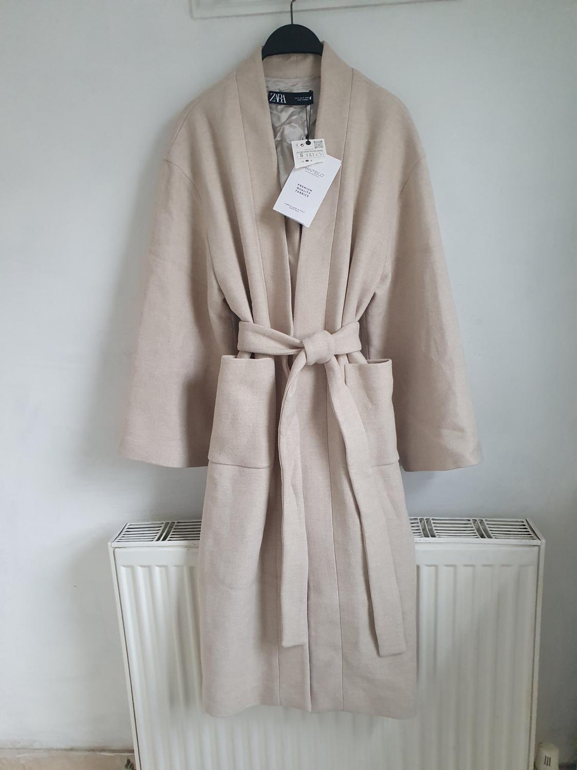 Beige coat hung on a black hanger over a white radiator. Visible labels with text: "Zara", "S", "Made in Morocco".