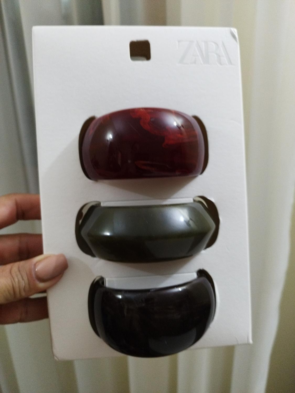 Three chunky bracelets in shades of red, green and black on a white stand with the Zara logo. Background of light-coloured curtains.
