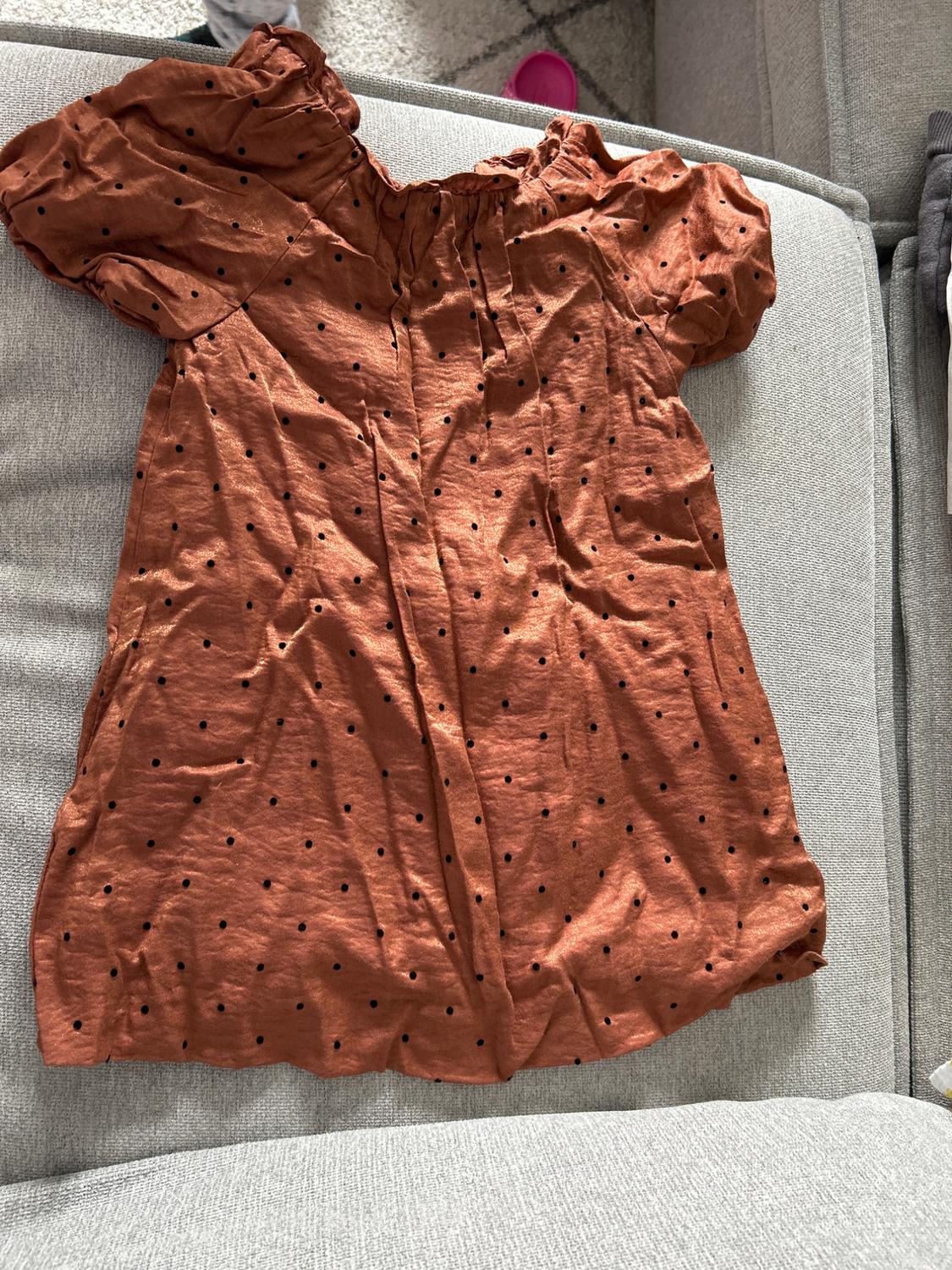 Brown dress with black polka dots on a grey sofa. Wrinkled fabric and short puff sleeves. Background with rug and cushion.